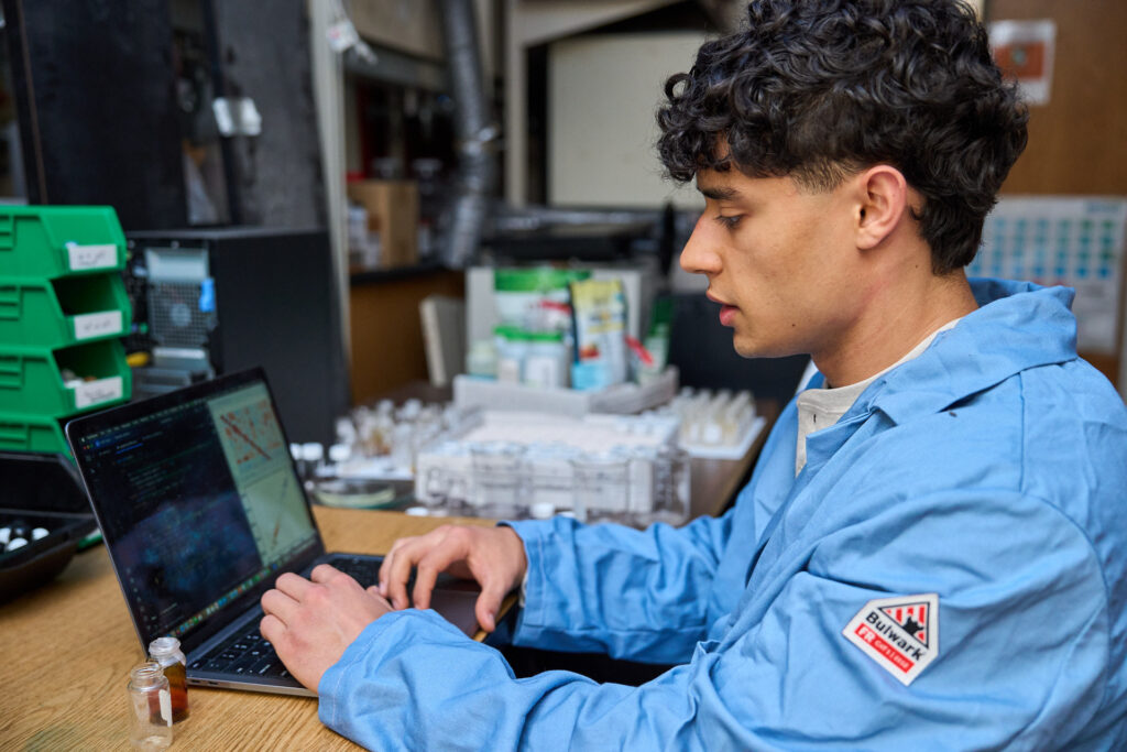 A man wearing a blue lab coat types something into a laptop in a science lab.