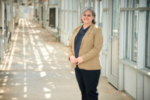 Μια γυναίκα που φορά ένα χακί σακάκι με σκούρο πουκάμισο και μαύρο παντελόνι στέκεται σε έναν μακρύ ανοιχτόχρωμο διάδρομο ενός θερμοκηπίου.