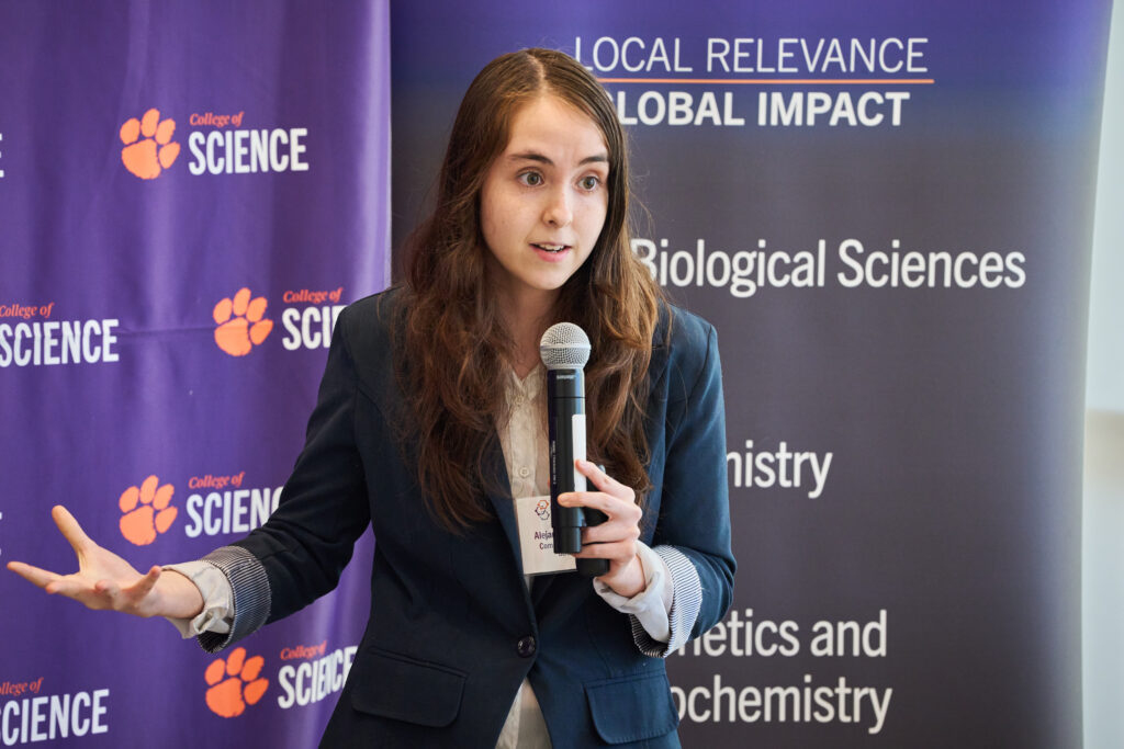 A woman wearing a black suit and holding a microphone talks to an audience during the Catalyst Competition. She's standing in front of a purple banner that has the College of Science logo and Tiger Paw multiple times.