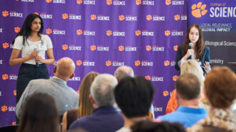 Two women stand in front of a step and repeat banner for the College of Science presenting to a crowd, which you can see in the foreground of the photo.