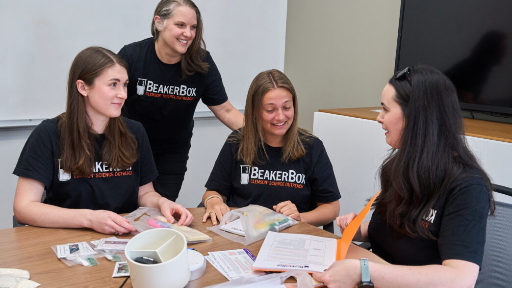 Three women students wearing black BeakerBox shirts assemble Beaker Box kits with science lessons and experiments that will be given to middle school science teachers while sitting at a table, while Kara Powder, an associate professor in the Department of Biological Sciences who is also dressed in a similar shirt, watches.