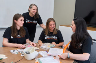 Three women students wearing black BeakerBox shirts assemble Beaker Box kits with science lessons and experiments that will be given to middle school science teachers while sitting at a table, while Kara Powder, an associate professor in the Department of Biological Sciences who is also dressed in a similar shirt, watches.