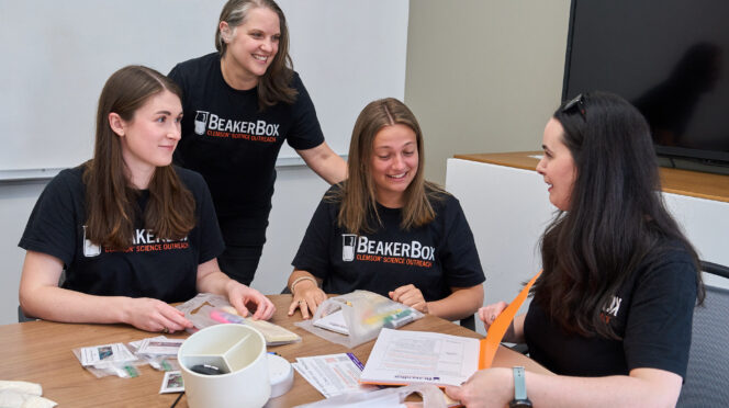 Three women students wearing black BeakerBox shirts assemble Beaker Box kits with science lessons and experiments that will be given to middle school science teachers while sitting at a table, while Kara Powder, an associate professor in the Department of Biological Sciences who is also dressed in a similar shirt, watches.