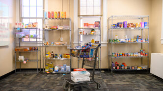 A shopping cart filled with pantry staples sits in front of four wire shelves in a food pantry.
