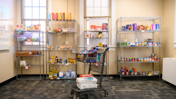 A shopping cart filled with pantry staples sits in front of four wire shelves in a food pantry.