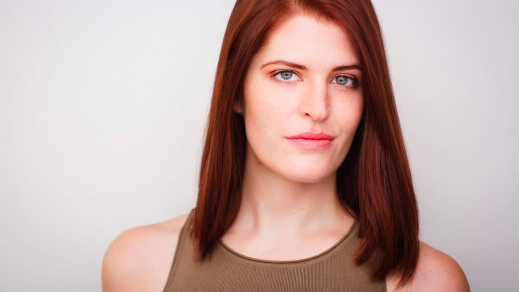 A young woman with shoulder-length red hair and a tan tank top looks at the camera.