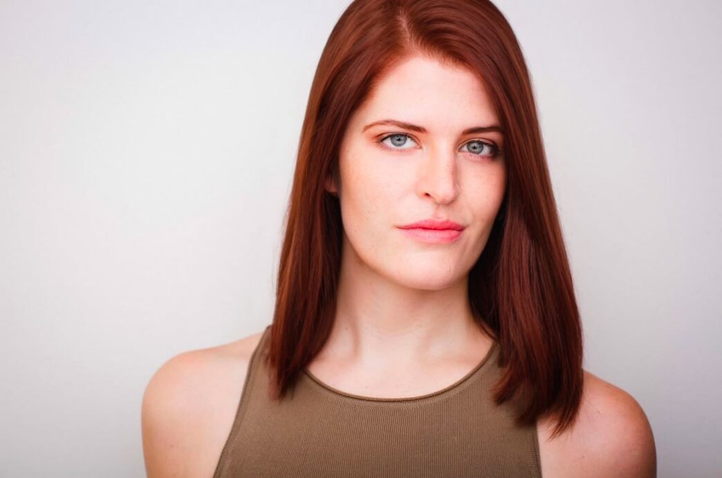 A young woman with shoulder-length red hair and a tan tank top looks at the camera.