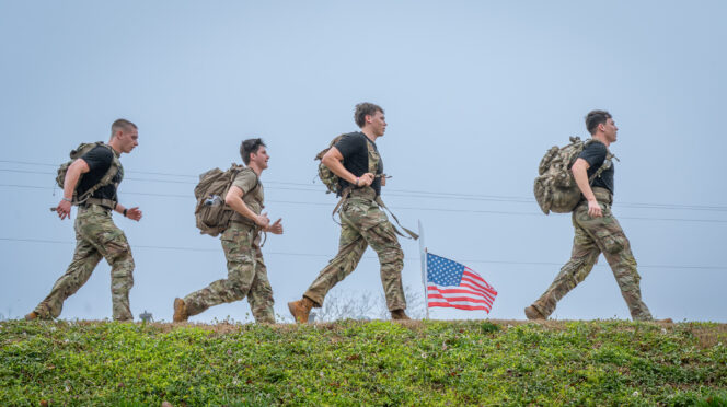 Cadets toting rucksacks walk on campus with an American flag in the background