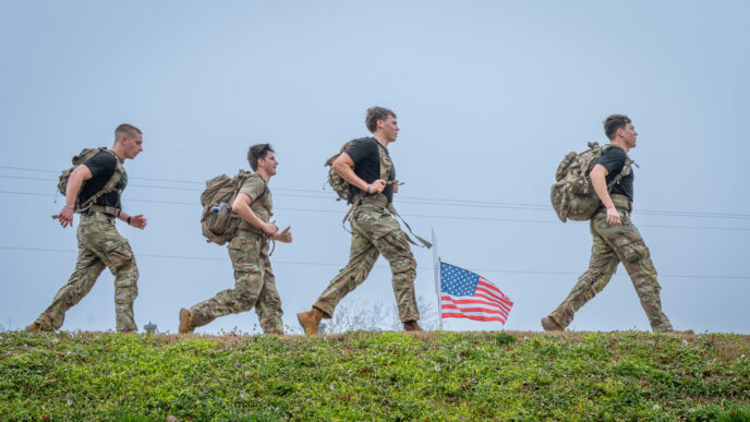 Cadets toting rucksacks walk on campus with an American flag in the background