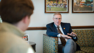 Doug Hallenbeck, Vice President for Student Affairs, sits on a couch in a suit speaking to another individual