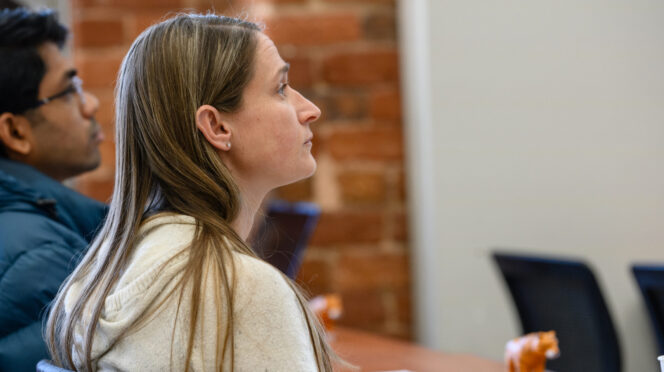 A woman in profile listens attentively in a classroom setting, with another person blurred in the background.