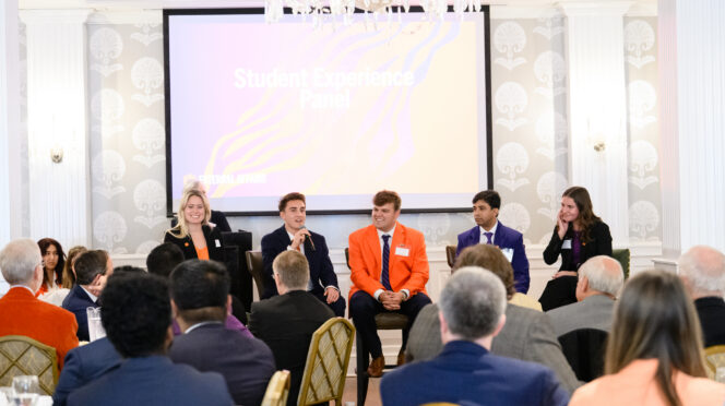 Five students sitting in chairs speaking to an audience in a ballroom, in front of a digital screen