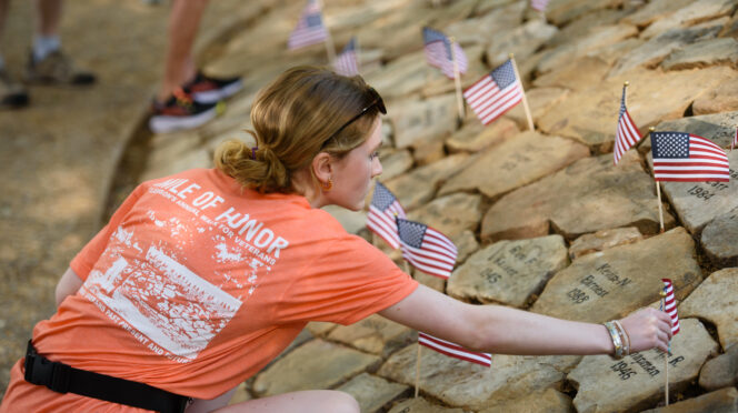 A female wearing an orange t-shirt places a miniature American flag into a stone barrow