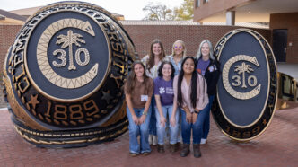 Six female Darla Moore Scholars, posing between two giant Clemson University class ring replicas, two of them are wearing vet med t-shirts