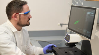 A man wearing goggles and a white lab coat and gloves looks at a computer screen showing an organism that is being looked at under a microscope
