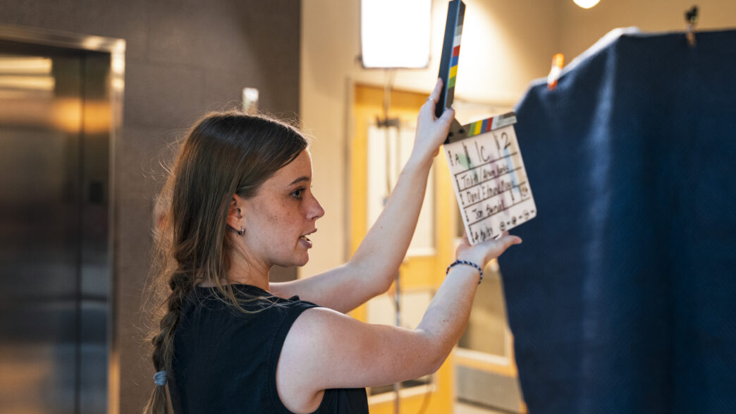 A young woman wearing a black tank top holds a movie clapperboard in the air, preparing to close it.
