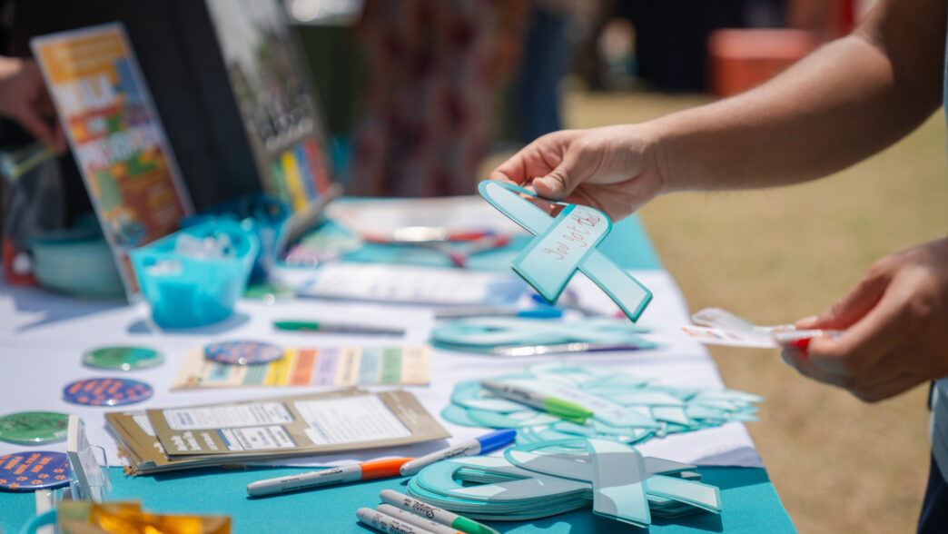 A person holds a teal awareness ribbon-shaped cutout with a handwritten message at a campus resource table filled with markers, pamphlets and craft materials during an outdoor event.