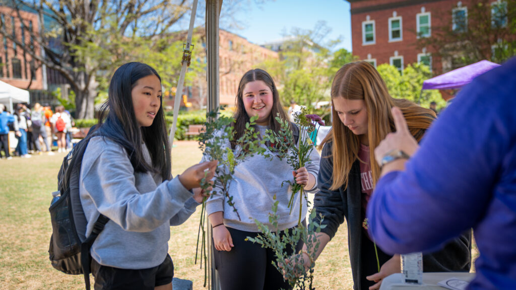 Three students participate in a hands-on activity at Yes Fest, holding and arranging flowers and greenery at a table outdoors, with campus buildings and other attendees visible in the background.