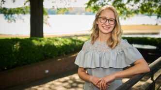 A woman with blonde hair and glasses wearing a dress leans up against a railing near a lake.