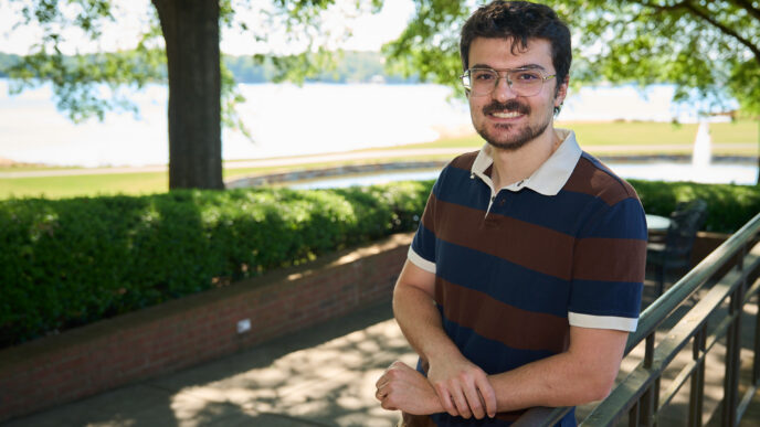 A man with dark hair and a beard and mustache wearing a short-sleeve rugby-style shirt leans up again a rail out by a lake