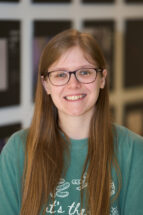 A headshot of a woman with long hair wearing glasses and a green t-shirt