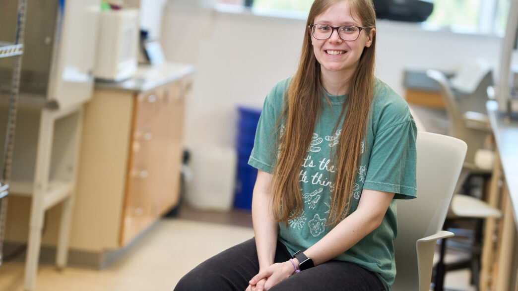 A woman with long hair and glasses wearing a green t-shirt sits in a chair in a science lab.