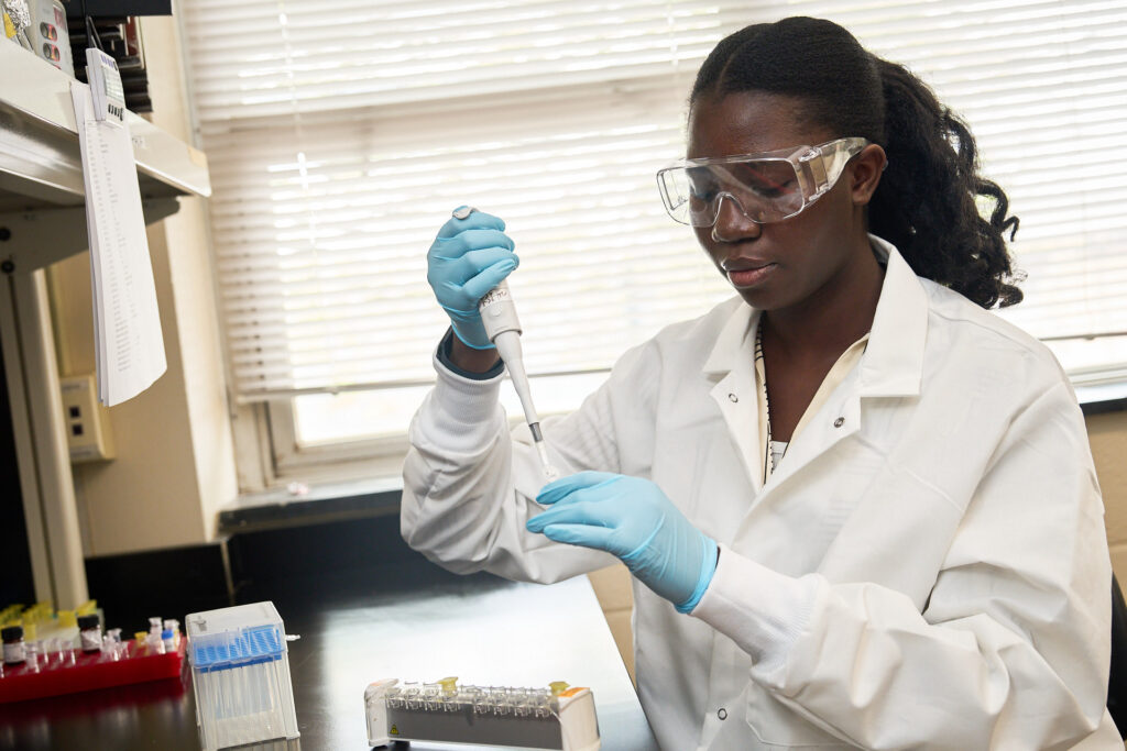 A woman gearing safety glasses, blue latex gloves and a white lab coat pipettes something into a container in a science lab.