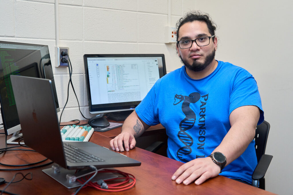 A man with dark hair, glasses and wearing a blue t-shirt sits at a desk with the computer monitor behind him and another in front of him.