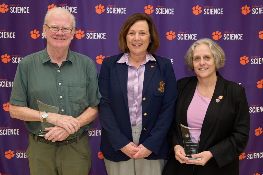 A man wearing a green shirt, a woman wearing a blue blazer and a woman wearing a black jacket and pink shirt stand in front of a banner with a bunch of tiger paws and College of Science logos. The two on the ends are holding acrylic trophies.