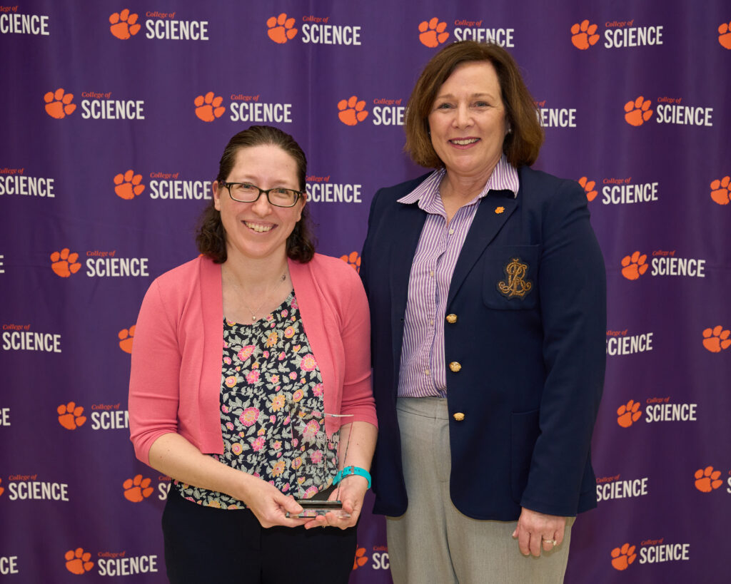 A woman wearing a pink sweater and holding an acrylic trophy stands next to a woman with a blue blazer. Both are standing in front of a banner with a lot of Tiger paws and College of Science logos.