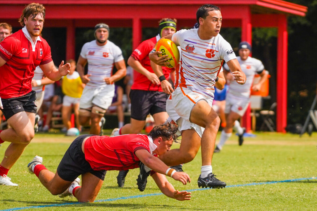 A rugby player in a white uniform is chased by several athletes in red uniforms and black shorts