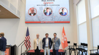 Three men wearing blazers stand on a stage flanked by a U.S. and Clemson Tiger Paw flag with a screen above them highlighting featured speakers for a real estate symposium.