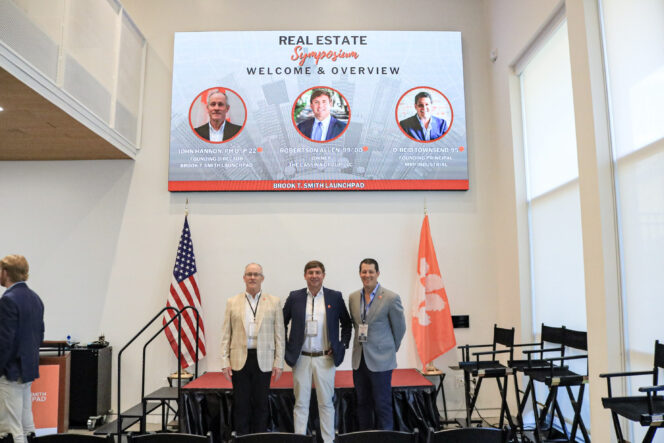 Three men wearing blazers stand on a stage flanked by a U.S. and Clemson Tiger Paw flag with a screen above them highlighting featured speakers for a real estate symposium.