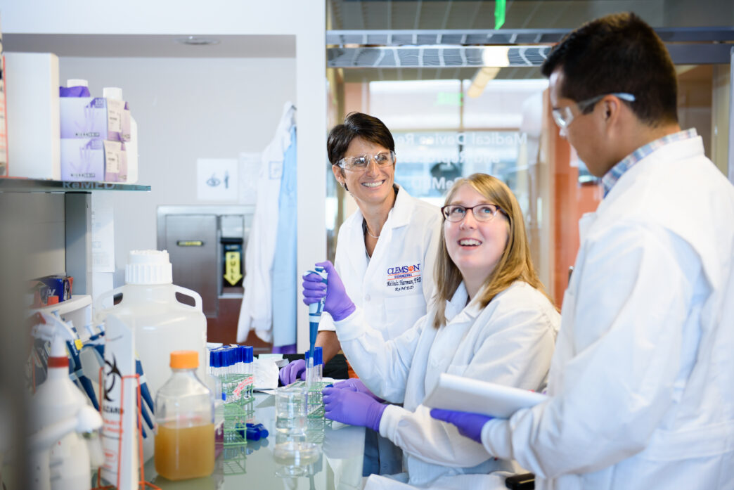 Three people wearing white lab coats and safety glasses work in a research laboratory.