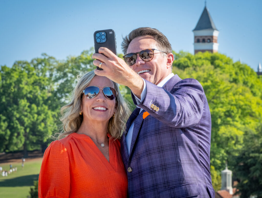 A man and woman stand next to each other and take a selfie with the Tillman clock tower visible behind them