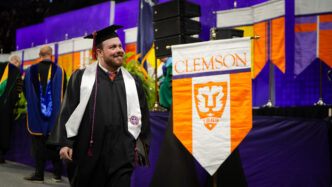 A student in graduation regalia (cap and gown) smiles with a Clemson University flag in the background