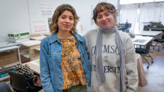 Two women, twins, stand with their arms around each other in an art studio