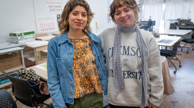 Two women, twins, stand with their arms around each other in an art studio