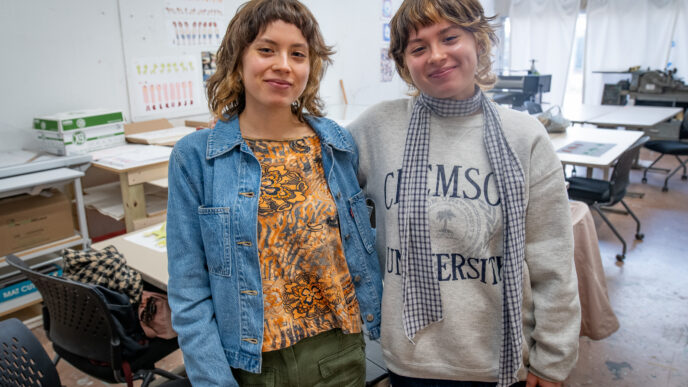 Two women, twins, stand with their arms around each other in an art studio
