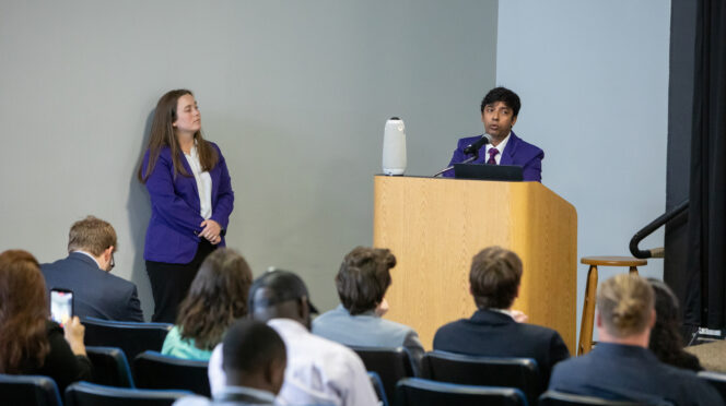 Individuals in purple blazers speak from a podium to an assembly