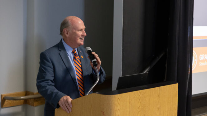 A male in a suit with Tiger Paw lapel and an orange tie speaks from a podium