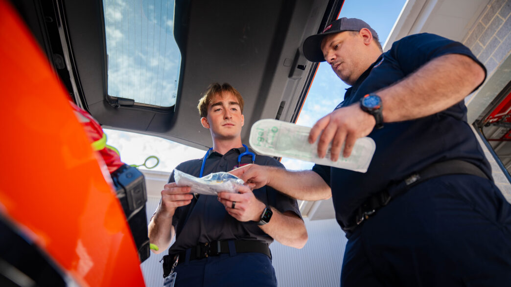 Two males look through EMS tools in the open trunk of a vehicle