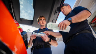 Two males look through EMS tools in the open trunk of a vehicle
