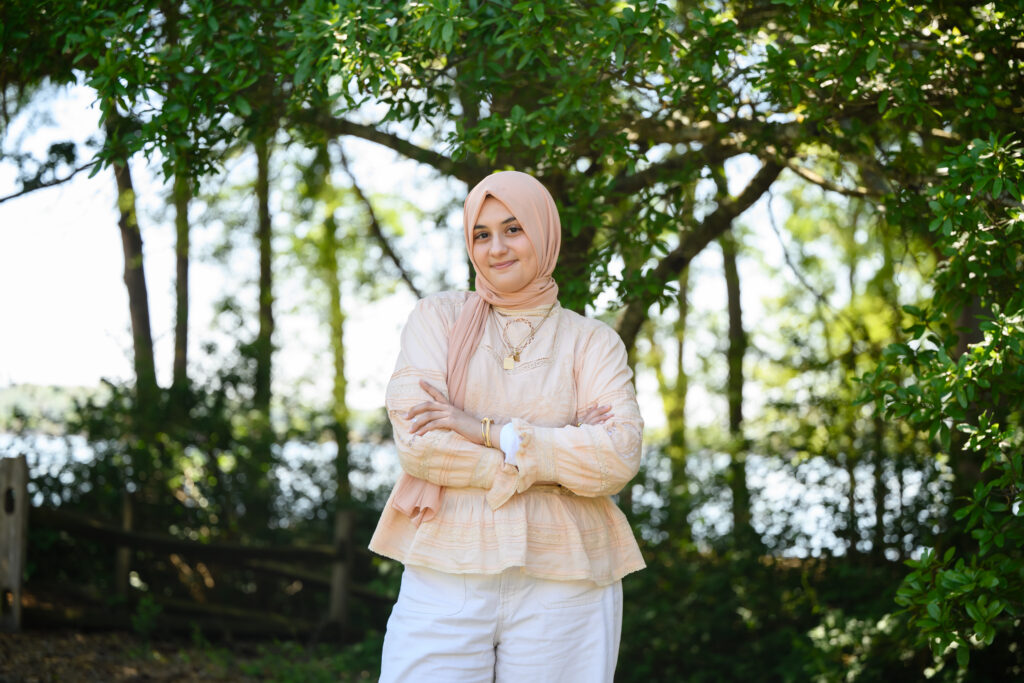 A young woman stands in front of greenery with her arms crossed. She is wearing white pants, a cream top and a light pink hijab.