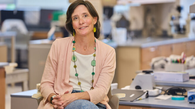 A woman with dark hair wearing a pale peach sweater and a white blouse with a green bead necklace and jeans sits near a lab table in a science lab with her legs crossed and hands in her lap.