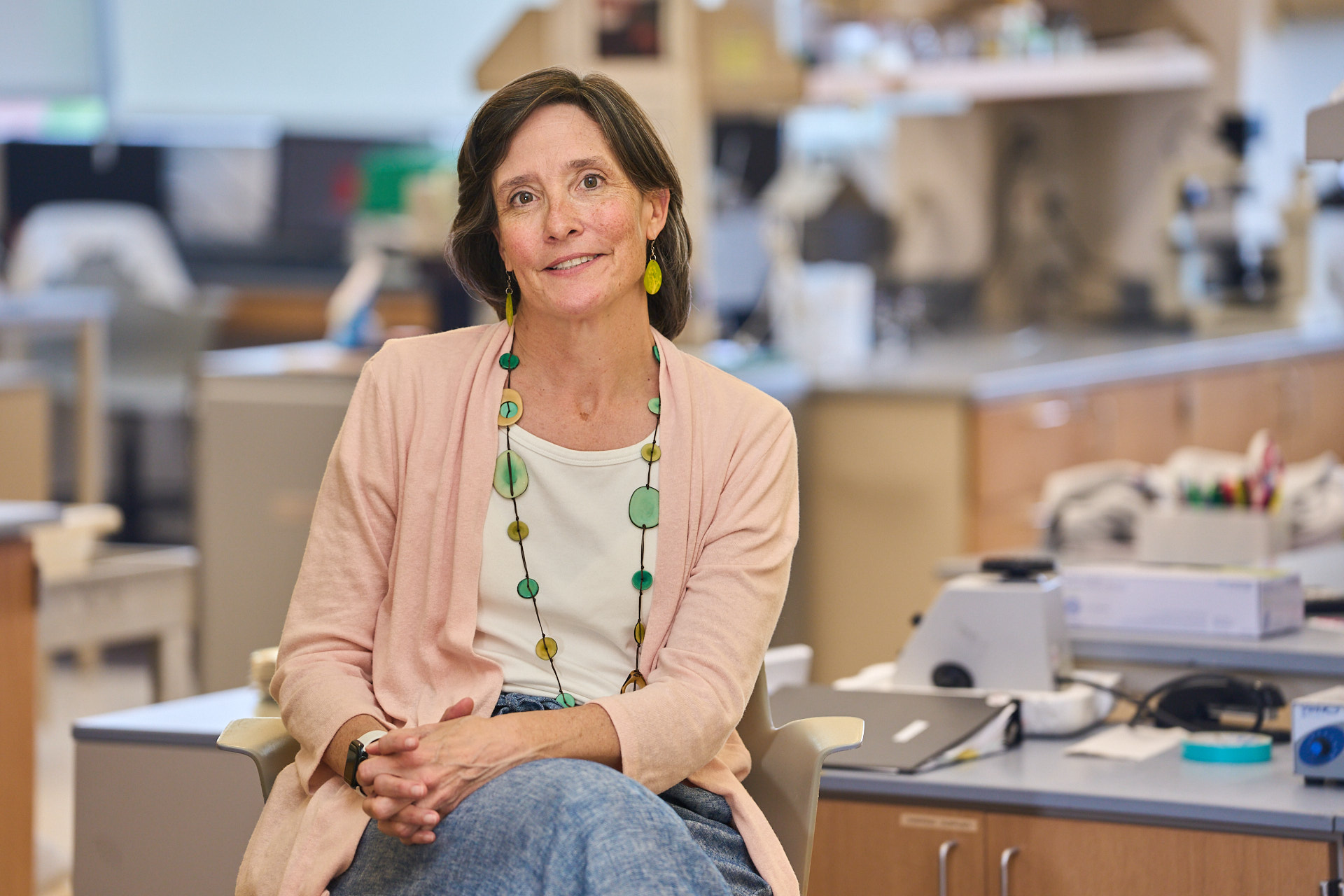 A woman with dark hair wearing a pale peach sweater and a white blouse with a green bead necklace and jeans sits near a lab table in a science lab with her legs crossed and hands in her lap.