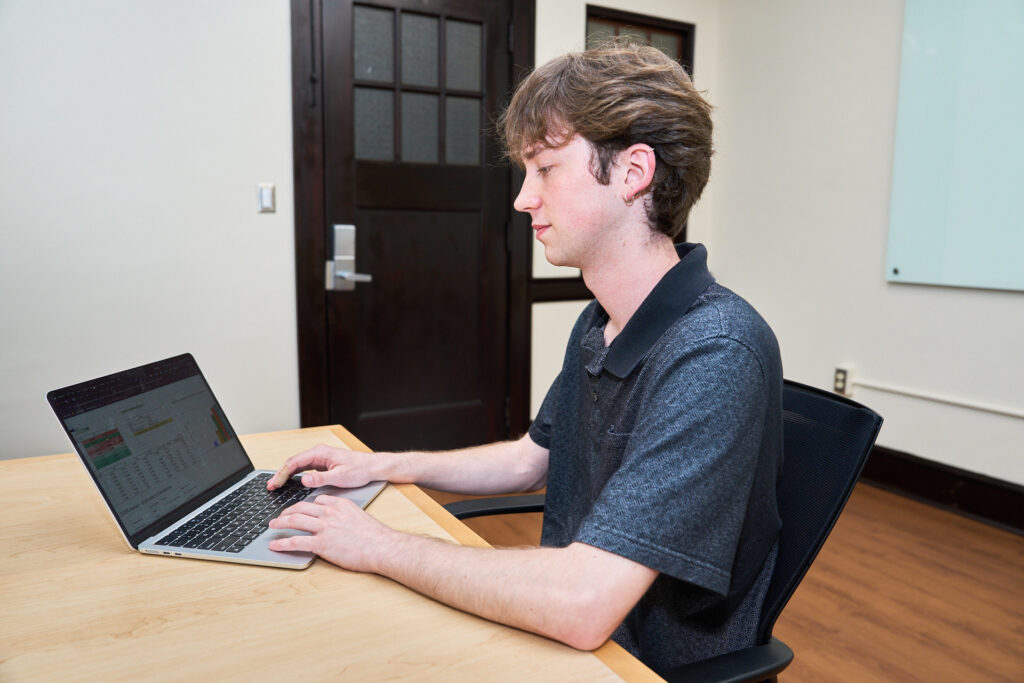 A man wearing a dark polo shirt sits at a desk looking at a laptop computer