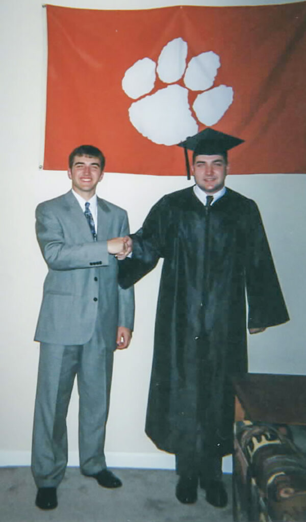Two men shake hands, one dressed in a suit and the other in graduation cap and gown, with a Clemson Tiger paw flag hanging on a wall above and behind them.
