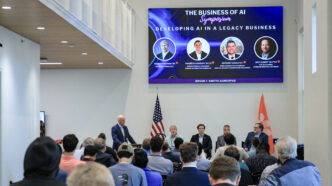 A room has several dozen people seated and looking at a stage with a panel of business people sitting, facing them. There are two flags on the stage (state of S.C. and U.S.) and a digital screen above them that reads "The business of AI."