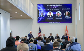A room has several dozen people seated and looking at a stage with a panel of business people sitting, facing them. There are two flags on the stage (state of S.C. and U.S.) and a digital screen above them that reads "The business of AI."